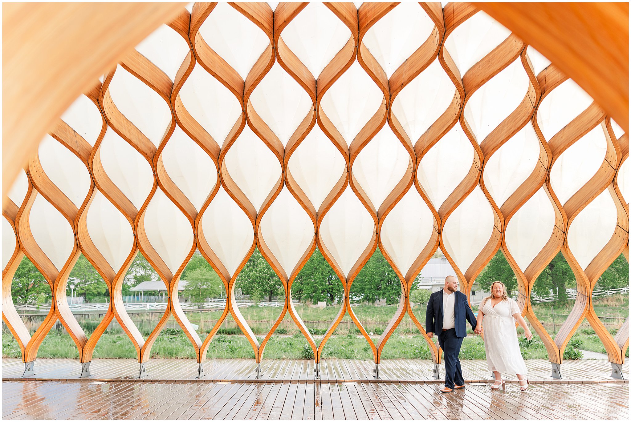 A couple walks along the Honeycomb structure in Lincoln Park Chicago, IL. The woman is wearing a white dress and holding the hand of a man wearing a navy suit.