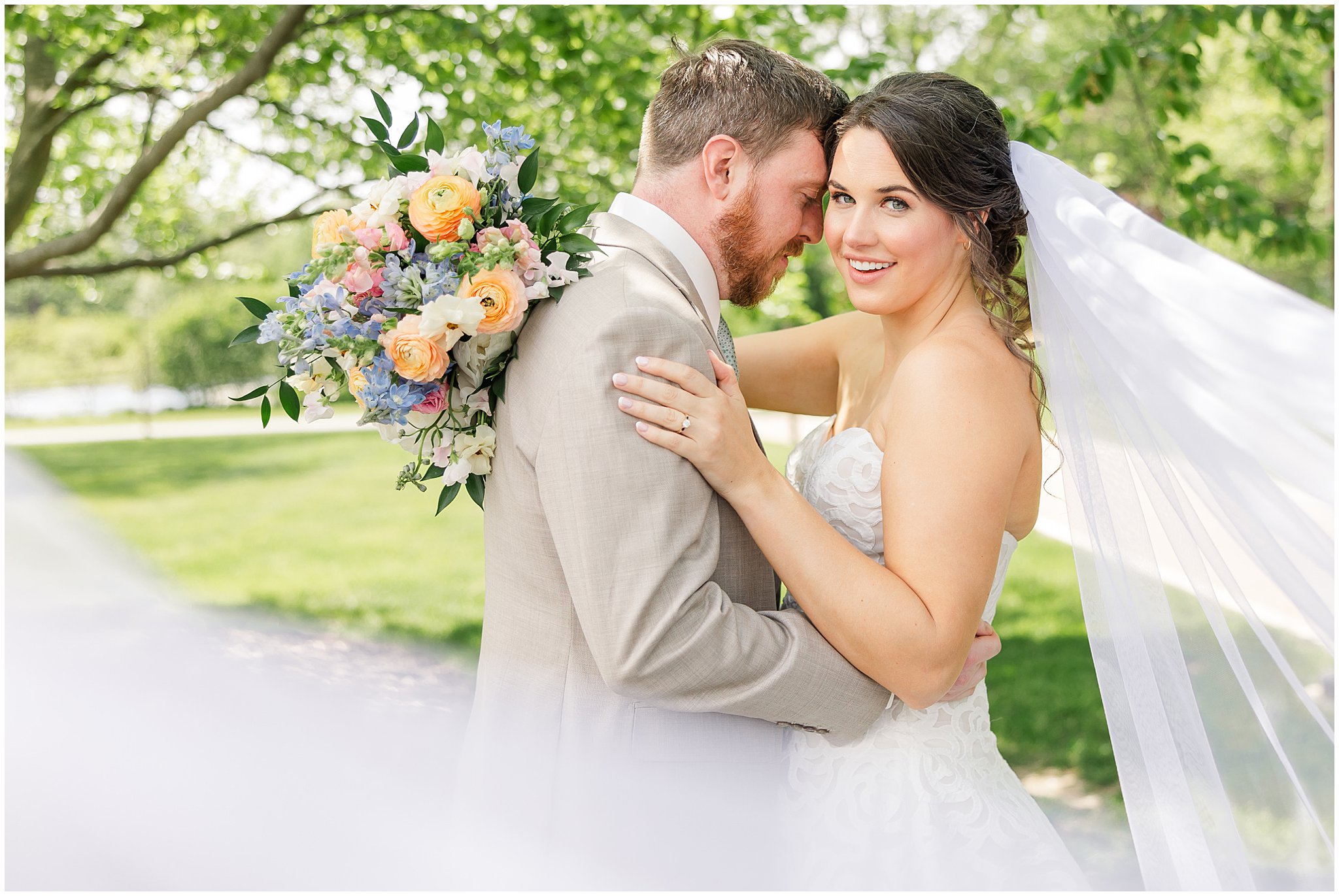 Bride and Groom on their wedding day, groom snuggles closely to the bride as she looks toward the camera with a big smile. The wedding veil flows through the bottom of the composition.