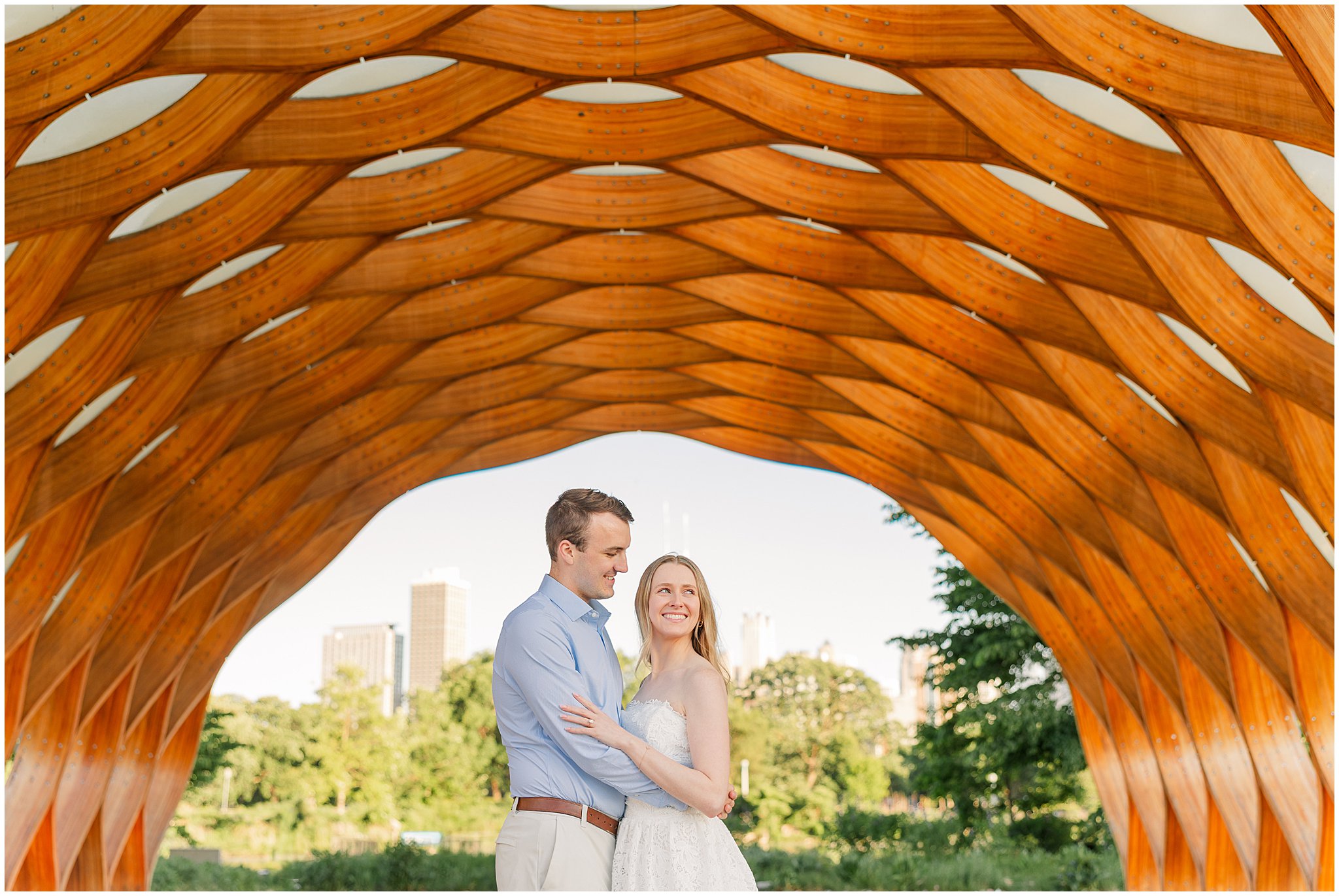 Engaged couple standing beneath the Honeycomb structure at Lincoln Park with the Chicago skyline in the distance.