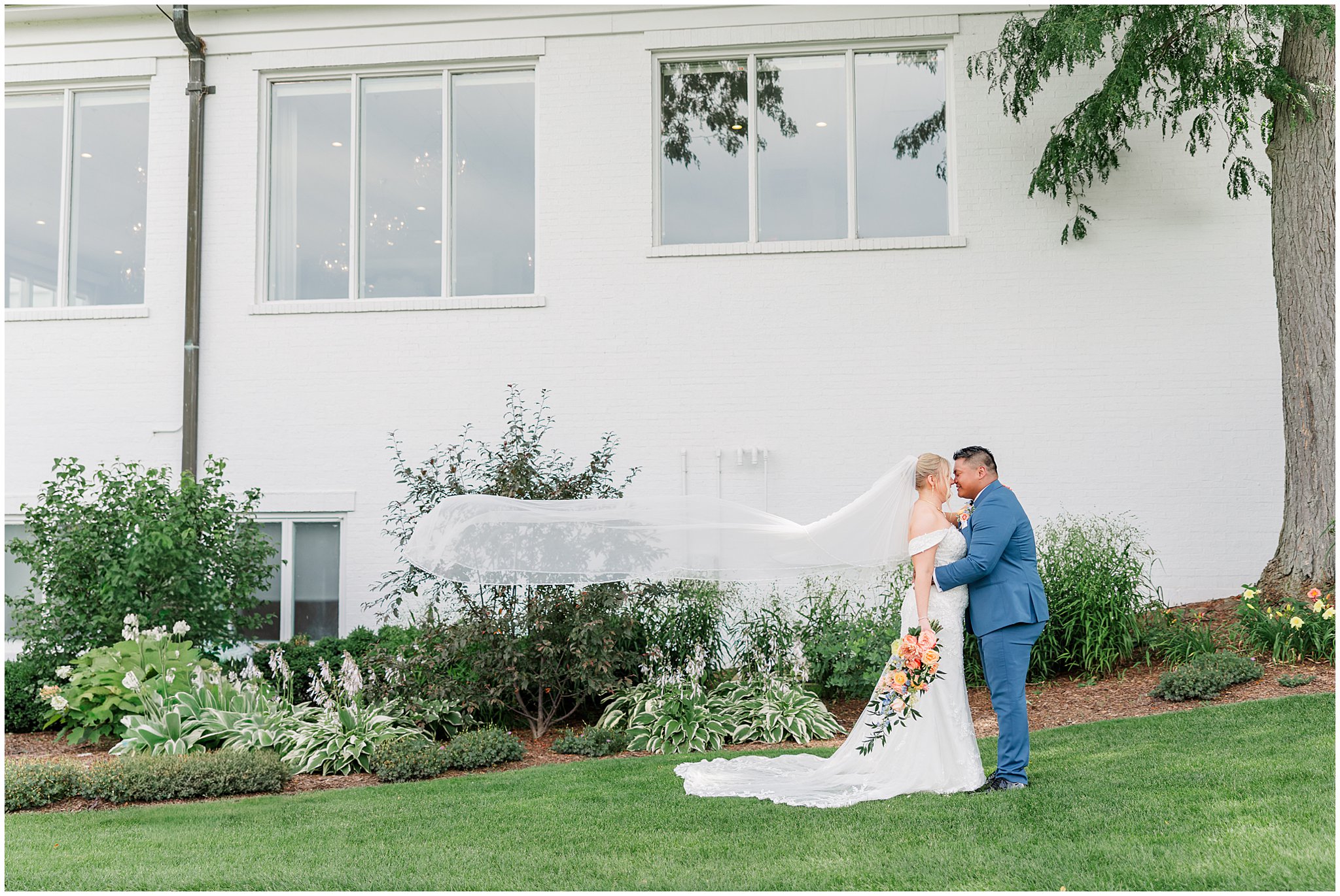 Bride & groom standing chest to chest in front of a building as the bride's veil flows in the air. She is holding her bouquet down at her side, as her groom snuggles in close. He is wearing a blue suit.